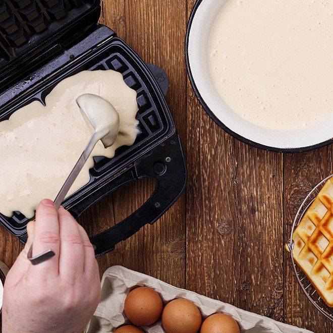 pâte à gaufre en train d'être coulée dans un gaufrier avec une boîte d'oeufs et un saladier rempli de la préparation de gaufres.